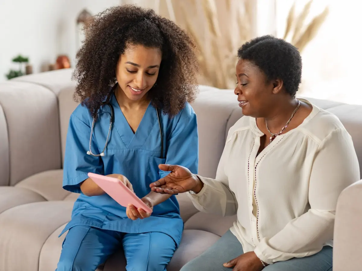 A carer in blue scrubs talking through a care plan with an older woman on her sofa