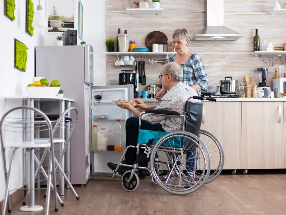 An older man using a wheelchair in his own kitchen while his carer prepares a meal