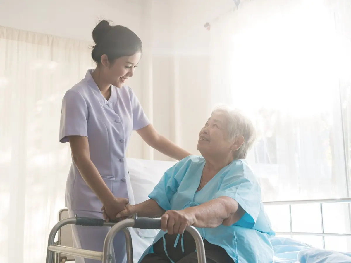 A carer helping an older woman up from her bed with her walking frame