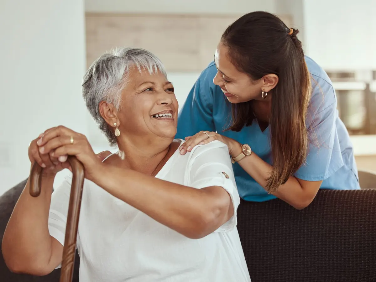 A smiling older woman holding her walking stick, with her carer’s hand on her shoulder